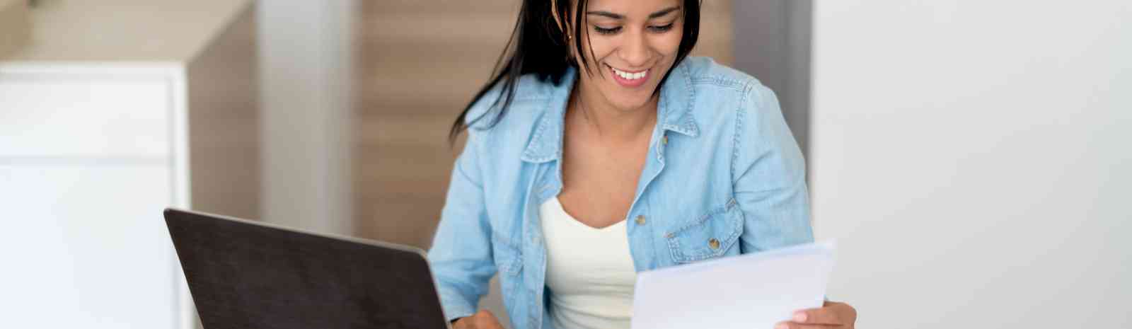 Young woman sitting with a laptop looking at a paper.